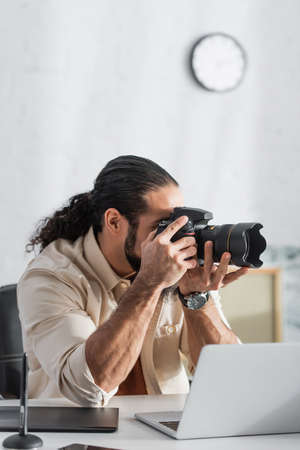 hispanic man taking photo on digital camera near blurred laptop at homeの写真素材