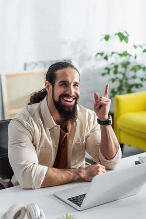 cheerful hispanic freelancer pointing up with finger while working near laptop at homeの写真素材