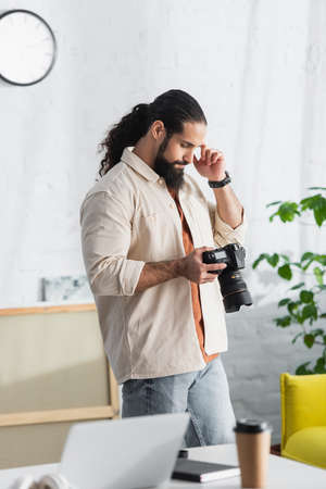 thoughtful hispanic photographer holding hand near head while looking at digital camera at homeの写真素材