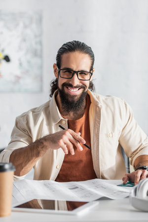 happy hispanic designer with pencil looking at camera near blurred blueprintの写真素材