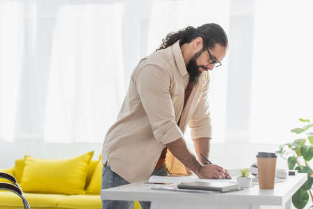 hispanic designer holding pencil while standing near work desk at home