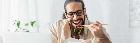 positive hispanic freelancer in eyeglasses eating chinese noodles near blurred laptop, bannerの写真素材
