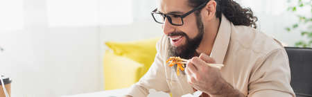 hispanic man in eyeglasses smiling while eating chinese noodles at home, banner