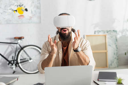 amazed hispanic freelancer in vr headset gesturing near blurred laptop at home