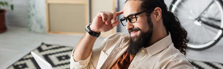 positive latin man in eyeglasses looking at digital tablet at home, banner