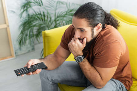 high angle view of thoughtful hispanic man with remote controller watching movie at home
