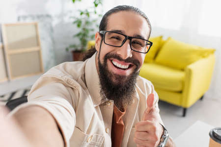cheerful latin man in eyeglasses looking ta camera and showing thumb up on blurred foregroundの写真素材