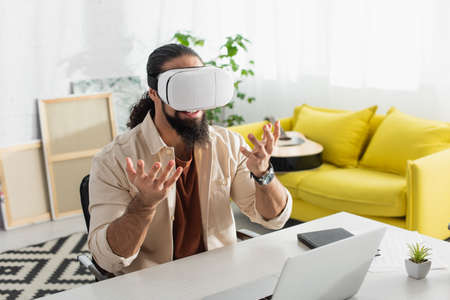 hispanic freelancer in vr headset gesturing near laptop at home