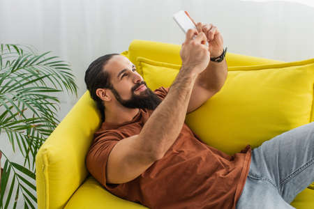 bearded latin man taking selfie on cellphone while lying on couch at home