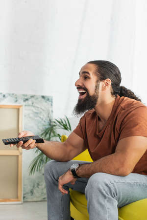 laughing latin man with remote controller watching comedy film at home