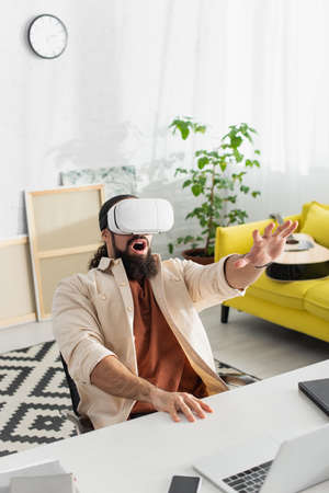 astonished hispanic man gesturing while gaming in vr headset in living room