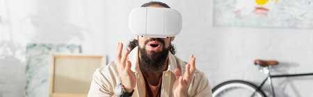 amazed hispanic man in vr headset showing wow gesture while gaming at home, banner