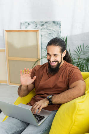cheerful latin man waving hand during video call on laptop near canvases on blurred background
