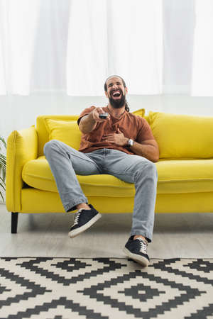 excited latin man with remote controller laughing on yellow sofa while watching tvの写真素材