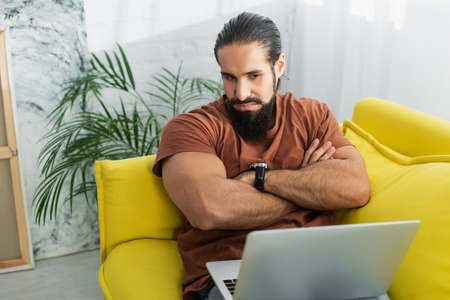serious hispanic man looking at laptop while sitting on couch at home