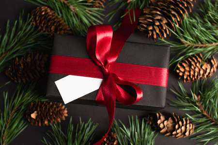 top view of wrapped gift box with red ribbon and blank card near fir branches and pine cones on blackの写真素材