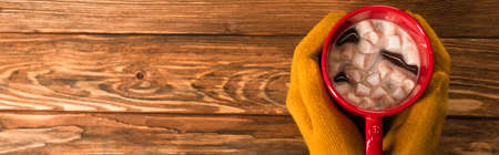 cropped view of person in yellow gloves holding cup of cocoa with marshmallows above wooden surface, bannerの写真素材