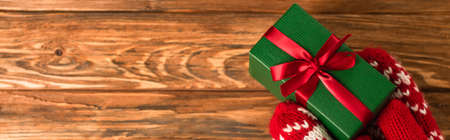 cropped view of person in red mittens holding wrapped gift box above wooden surface, banner