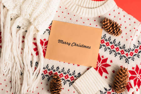 top view of greeting card with merry christmas lettering near pine cones on knitted sweaterの写真素材