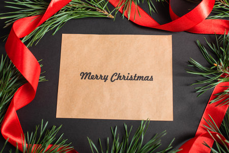 top view of greeting card with merry christmas lettering near fir branches and red ribbon on black