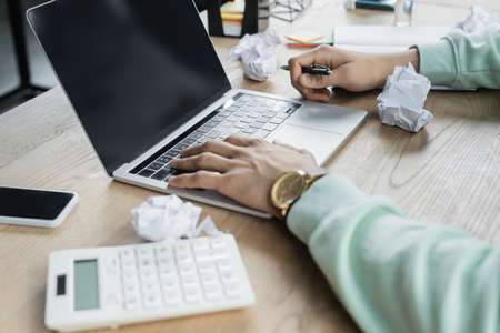 Cropped view of businessman with pen using laptop with blank screen near crumpled paper and blurred...