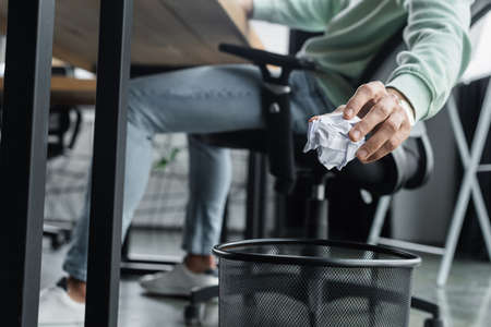 Cropped view of blurred businessman holding crumpled paper near trash can in officeの写真素材