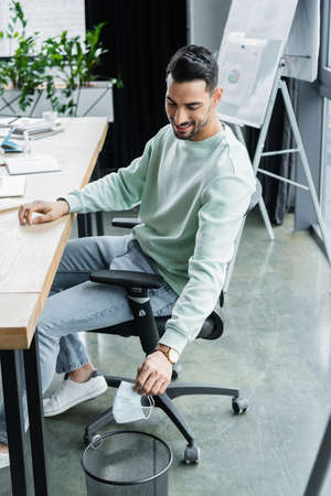 Positive muslim businessman holding medical mask near trash can in office