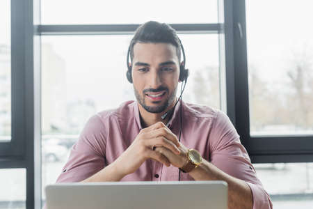 Smiling arabian businessman in headset having video call on blurred laptop in office