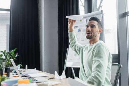 Smiling arabian businessman holding paper plane near laptop and calculator in officeの写真素材