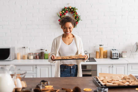 Smiling african american woman holding baked gingerbreads near ingredients in kitchenの写真素材