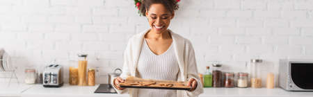 Positive african american woman holding baked christmas gingerbreads in kitchen, bannerの写真素材