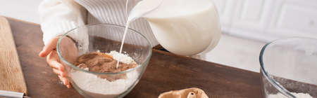 Cropped view of african american woman pouring milk in flour with cocoa while cooking in kitchen,...