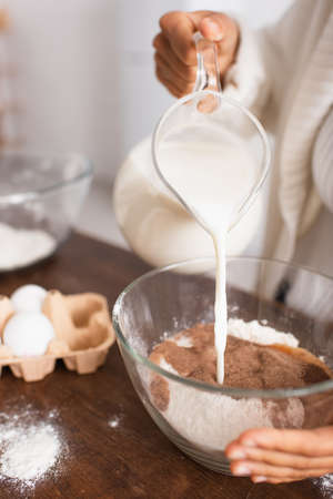 Cropped view of african american woman pouring milk in flour with cocoa near blurred eggs in kitchen