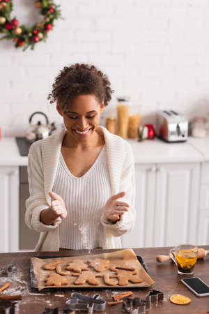 Smiling african american woman pouring flour on christmas cookies near smartphone and tea