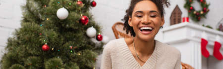 Cheerful african american woman looking at camera near blurred christmas tree at home, bannerの写真素材