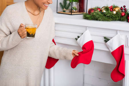 Cropped view of smiling african american woman with tea touching christmas stocking on fireplace at homeの写真素材