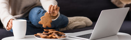 Cropped view of african american woman with cup taking christmas gingerbreads near laptop at home,...