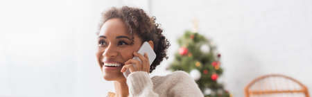 Smiling african american woman talking on smartphone near blurred christmas tree at home, banner