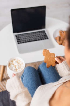 Christmas cookie and cup of cocoa with marshmallows in hands of african american woman near blurred laptopの写真素材