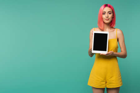 pleased young woman with pink dyed hair holding digital tablet with blank screen isolated on blueの写真素材