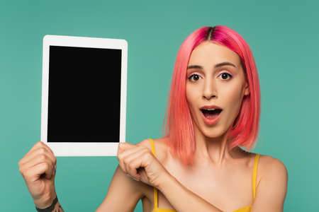 amazed young woman with pink dyed hair holding digital tablet with blank screen isolated on blueの写真素材