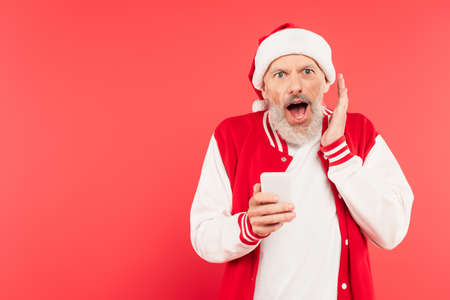 surprised middle aged man in santa hat holding smartphone isolated on red