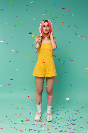 full length of happy young woman with pink dyed hair in wireless headphones near colorful confetti...