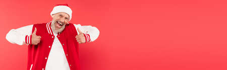 excited middle aged man in santa hat showing thumbs up isolated on red, banner