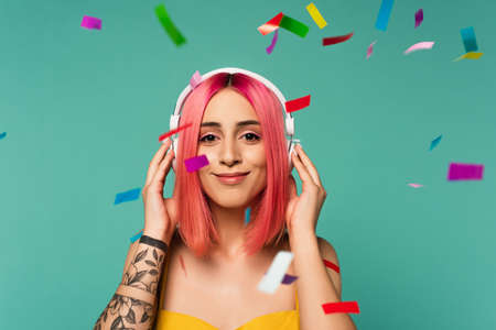 happy young woman with pink dyed hair in wireless headphones near colorful confetti on blueの写真素材