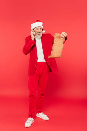 full length of positive mature man in santa hat reading letter on craft paper while talking on smartphone on redの写真素材