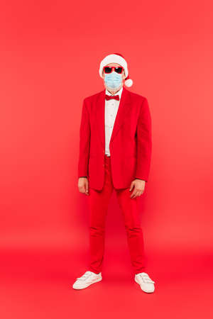 full length of mature man in santa hat, medical mask and sunglasses standing on redの写真素材