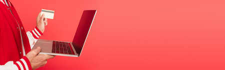 cropped view of middle aged man holding laptop and credit card isolated on red, bannerの写真素材