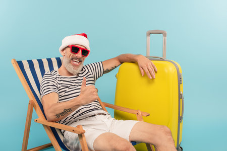 happy mature man in santa hat and sunglasses sitting on deck chair near luggage and showing thumb up isolated on blueの写真素材