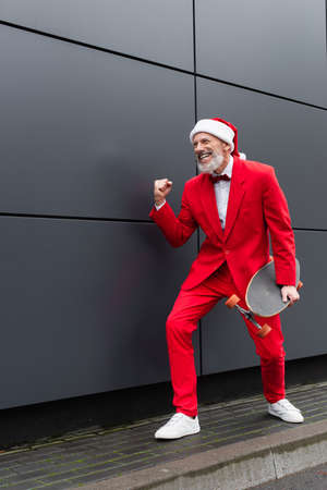 happy mature man in santa hat holding skateboard while standing near grey wallの写真素材